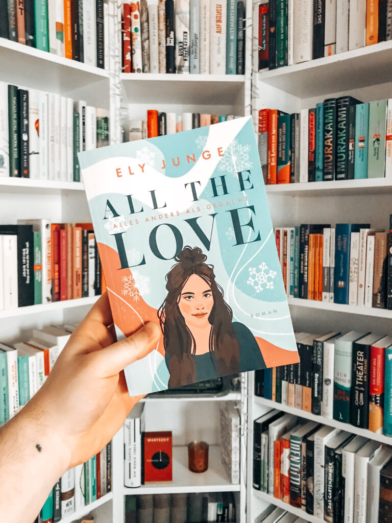 Das Taschenbuch von All the Love – Alles anders als gedacht von Ely Junge wird vor ein gut gefülltes Bücherregal gehalten